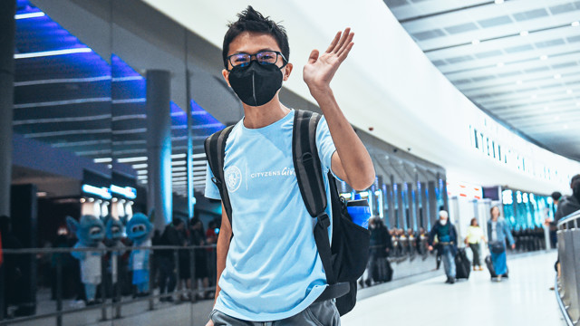 A person waving at the airport wearing a Cityzens Giving shirt and backpack, with Manchester City mascots in the background.