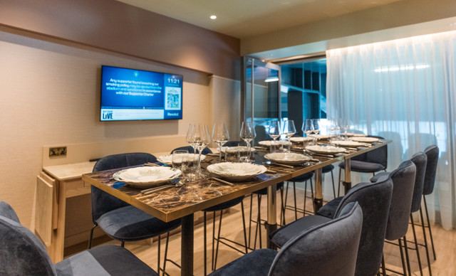High table dining setup with plates and glasses, surrounded by tall chairs. A screen displays information about a smoking policy, mentioning 'Revolut' and 'network LIVE'. Luxurious and modern interior with curtains.