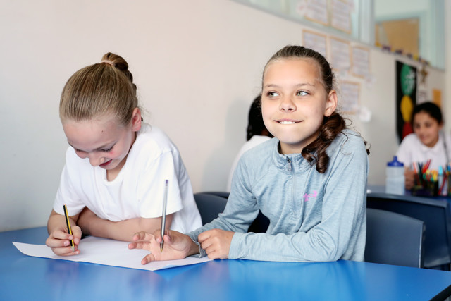 Two children, whose faces are blurred, are sitting at a blue table in a classroom. They appear to be engaged in writing or drawing on a piece of paper using pencils. In the background, other students are visible sitting at desks.