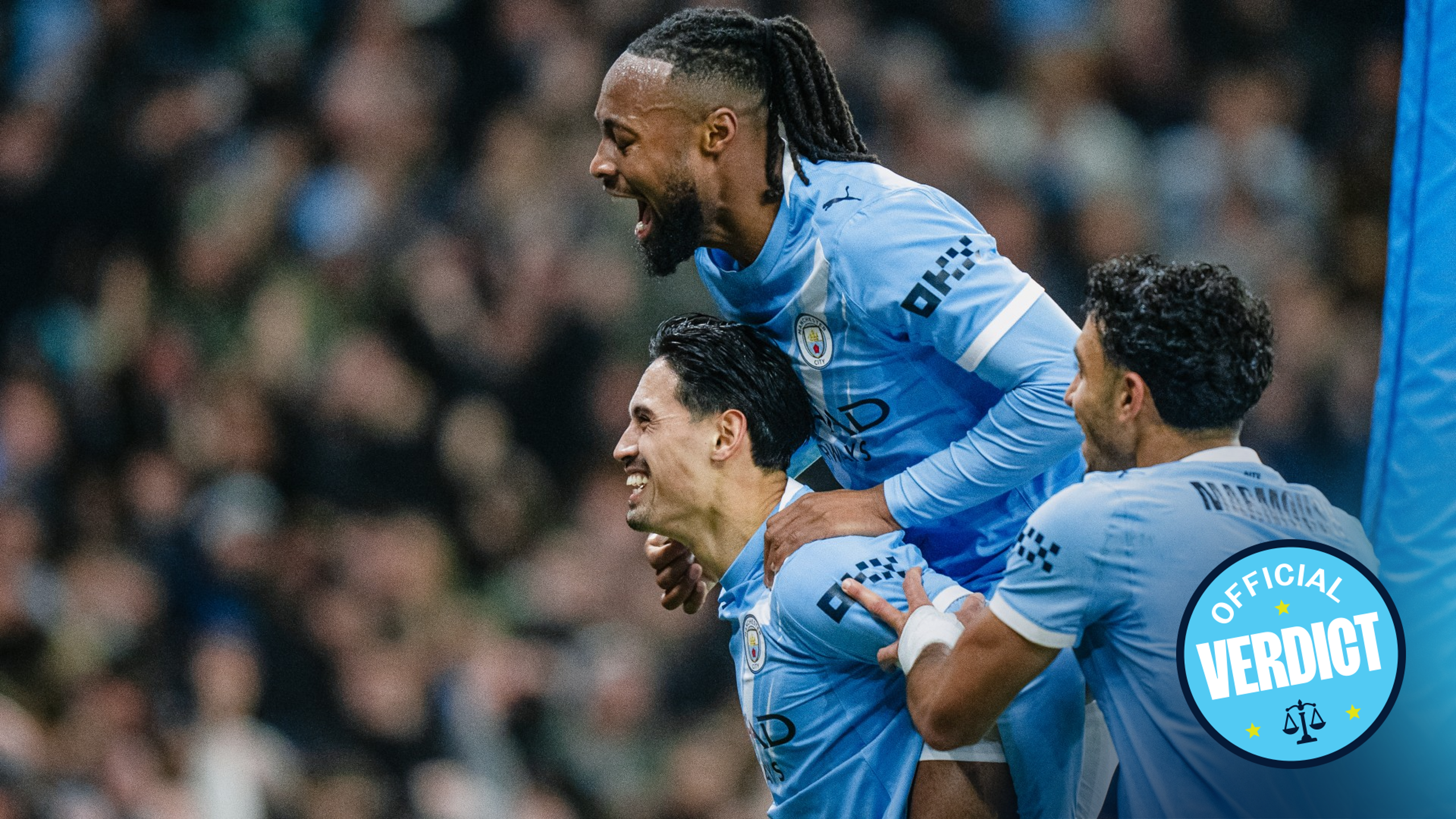 Manchester City players wearing light blue jerseys celebrate a goal during a match. The jersey features the Manchester City crest.