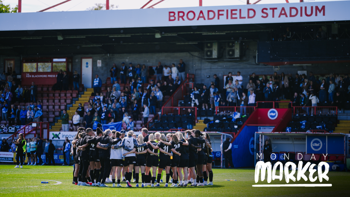 Soccer team huddle on the field at Broadfield Stadium with spectators in the stands. The words 'Monday Marker' are seen overlaying the image.