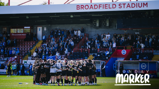 Soccer team huddle on the field at Broadfield Stadium with spectators in the stands. The words 'Monday Marker' are seen overlaying the image.