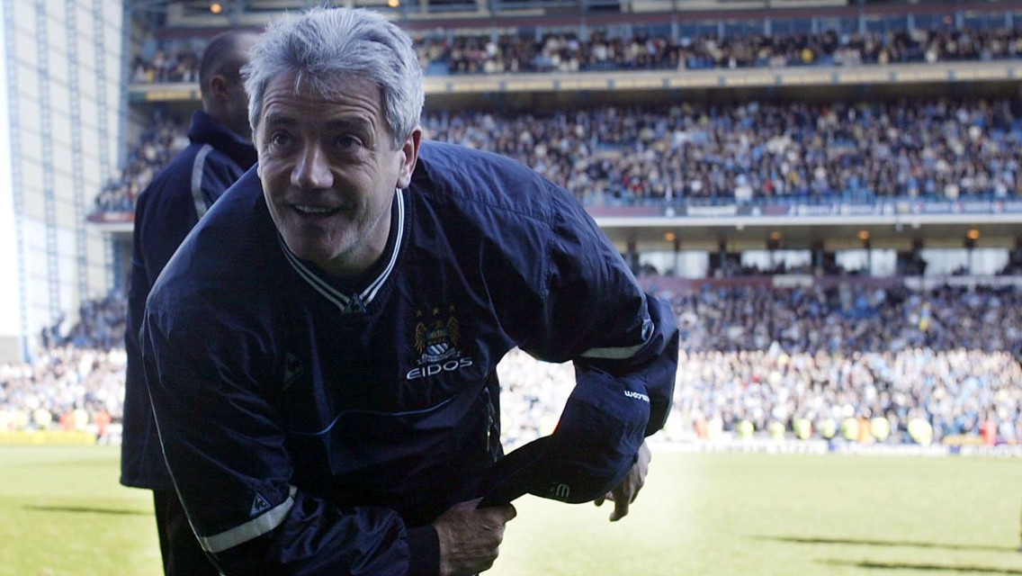 A coach wearing a Manchester City jacket stands on the field of a stadium filled with spectators.