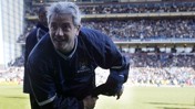 A coach wearing a Manchester City jacket stands on the field of a stadium filled with spectators.