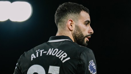 A football player seen from the back, wearing a jersey with the name 'Aït-Nouri' and Premier League logo, under bright stadium lights.