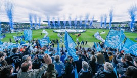 A vibrant crowd of Manchester City fans waving blue flags during a match at Joie Stadium with fireworks display in the background.