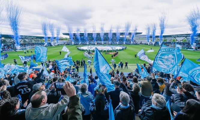 A vibrant crowd of Manchester City fans waving blue flags during a match at Joie Stadium with fireworks display in the background.