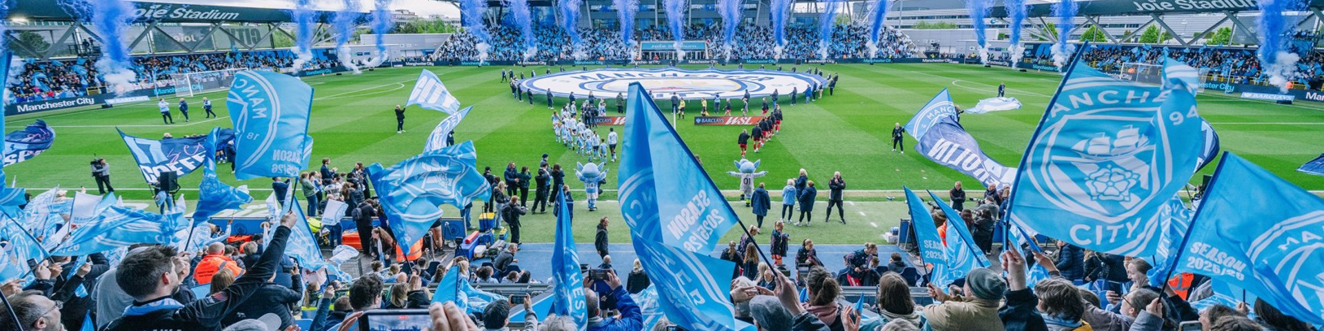 A vibrant crowd of Manchester City fans waving blue flags during a match at Joie Stadium with fireworks display in the background.