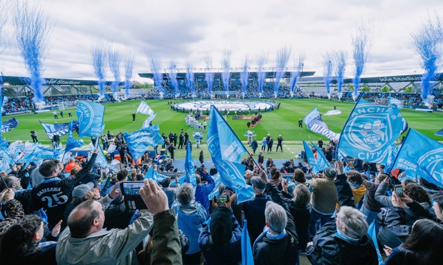 A vibrant crowd of Manchester City fans waving blue flags during a match at Joie Stadium with fireworks display in the background.