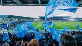 Fans waving Manchester City flags during a match at the Academy Stadium.