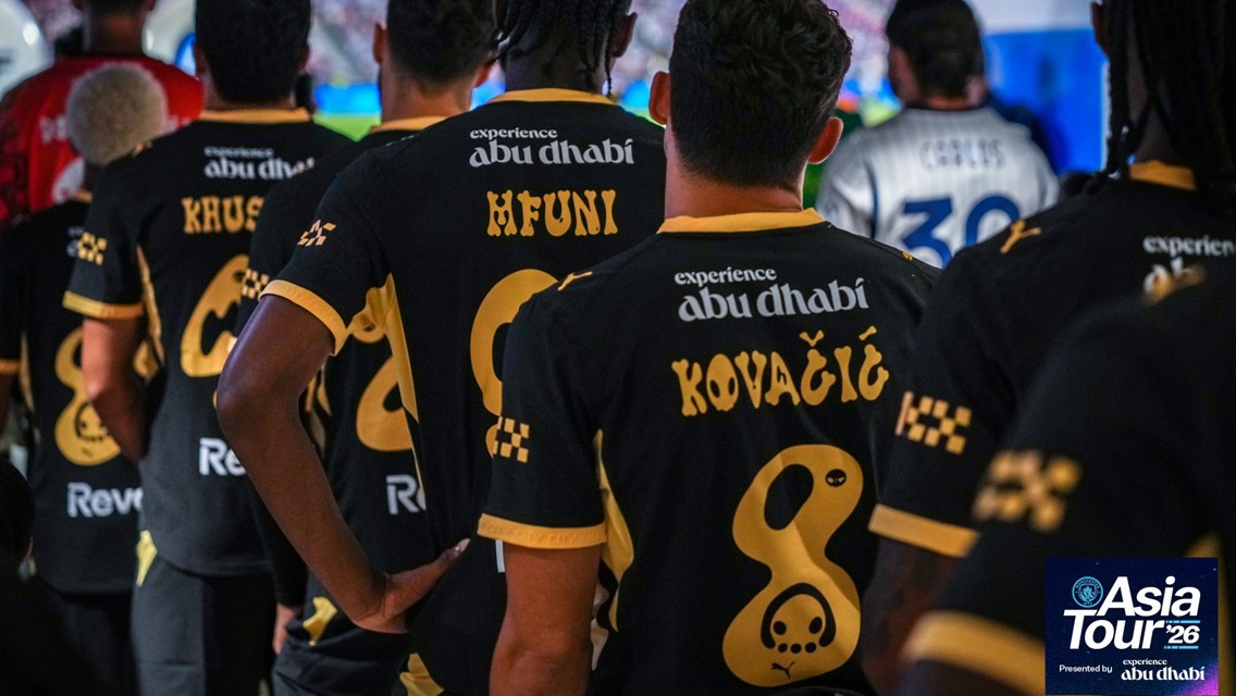 Football team wearing black and gold jerseys with player names in a tunnel. The jerseys promote experience Abu Dhabi. Asia Tour '26 mentioned in the corner.