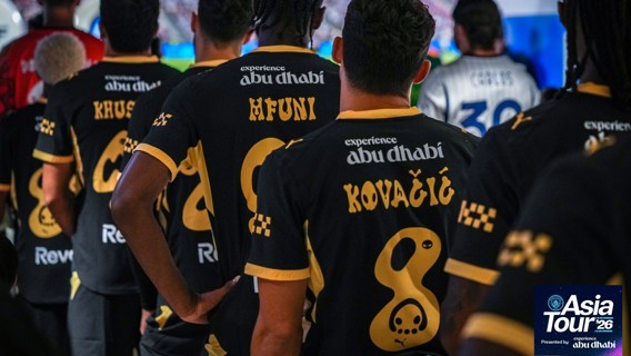 Football team wearing black and gold jerseys with player names in a tunnel. The jerseys promote experience Abu Dhabi. Asia Tour '26 mentioned in the corner.