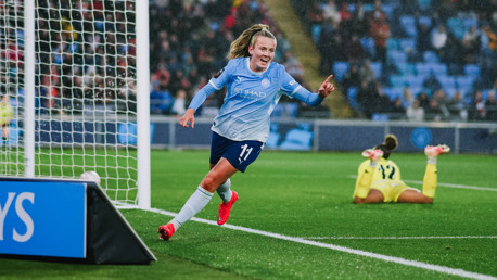 A soccer player from Manchester City Women's team celebrates a goal during a match, wearing a light blue jersey with the number 11. The goalkeeper is visible on the ground in the background.