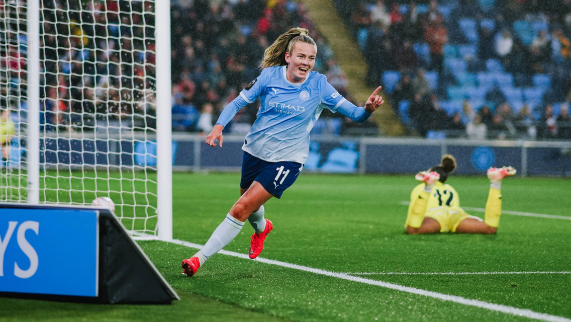 A soccer player from Manchester City Women's team celebrates a goal during a match, wearing a light blue jersey with the number 11. The goalkeeper is visible on the ground in the background.