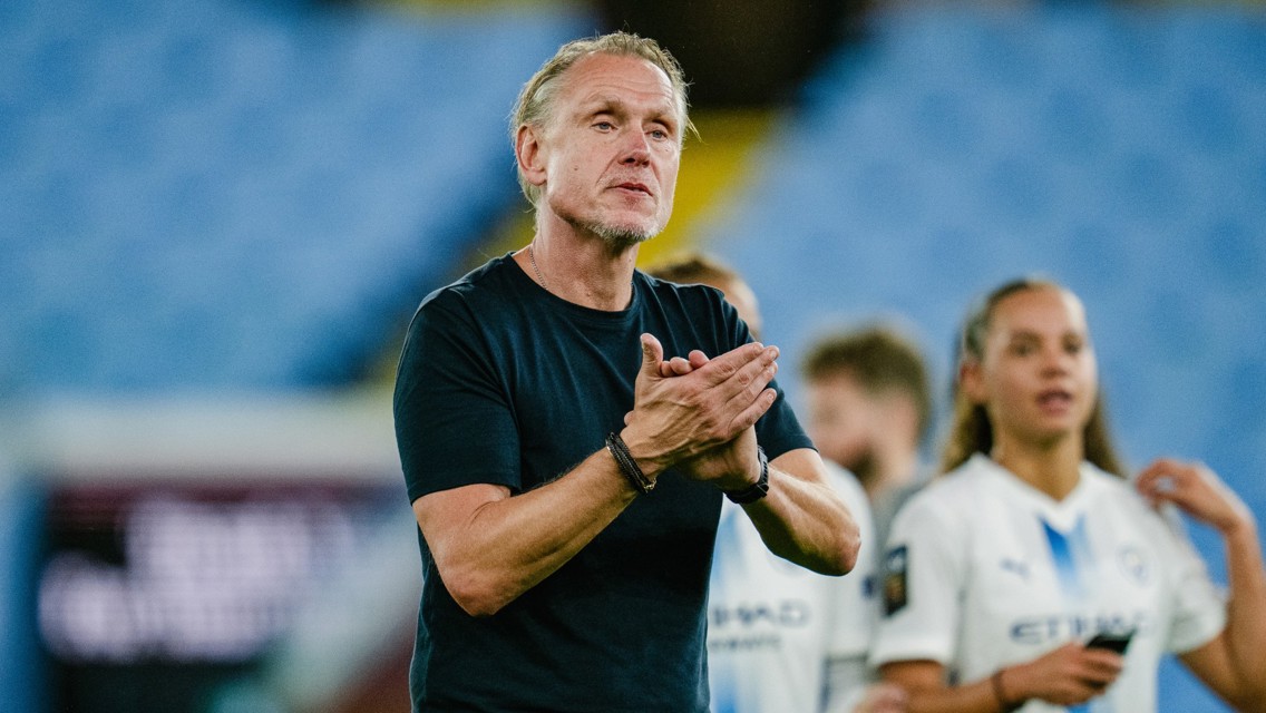 A person in a black shirt is clapping, with Manchester City Women's team members in the background at a stadium.