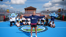 A person in a 'City in the Community' hoodie is surrounded by children and two blue Manchester City mascots on a blue sports court.
