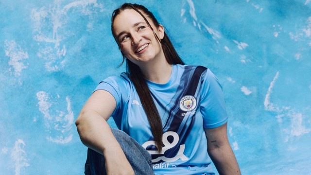 A person wearing a Manchester City esports jersey sits against a blue background. The jersey has the club crest and sponsor logos visible.