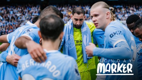 Manchester City players huddle together on the pitch with a towel draped over one's shoulder, wearing blue jerseys. 'Monday Marker' text is visible.
