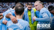 Manchester City players huddle together on the pitch with a towel draped over one's shoulder, wearing blue jerseys. 'Monday Marker' text is visible.