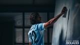 A person in a Manchester City uniform with 'Bouaddi' and number '32' spray painting a wall indoors. Text reads 'Welcome to City Ayyoub Bouaddi'.