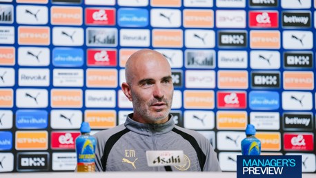 A man speaking at a press conference, wearing a Manchester City jacket, with sponsor logos in the background.