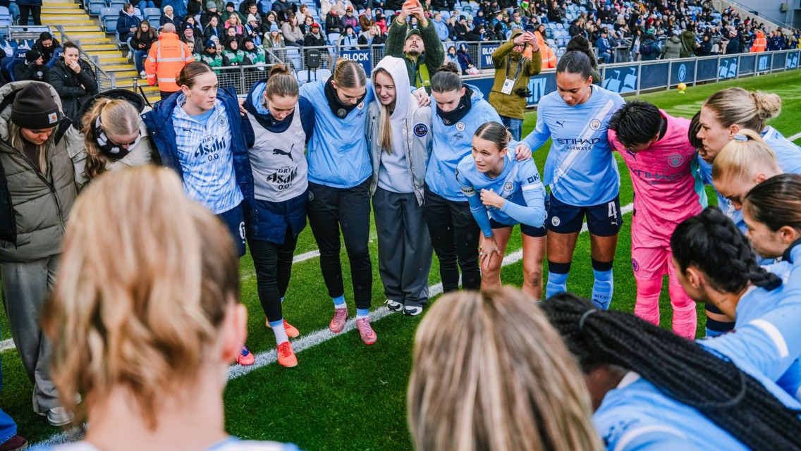The image shows the Manchester City women's football team huddled together on a pitch before a game, wearing their team kits. They are surrounded by spectators in a stadium.
