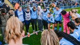 The image shows the Manchester City women's football team huddled together on a pitch before a game, wearing their team kits. They are surrounded by spectators in a stadium.