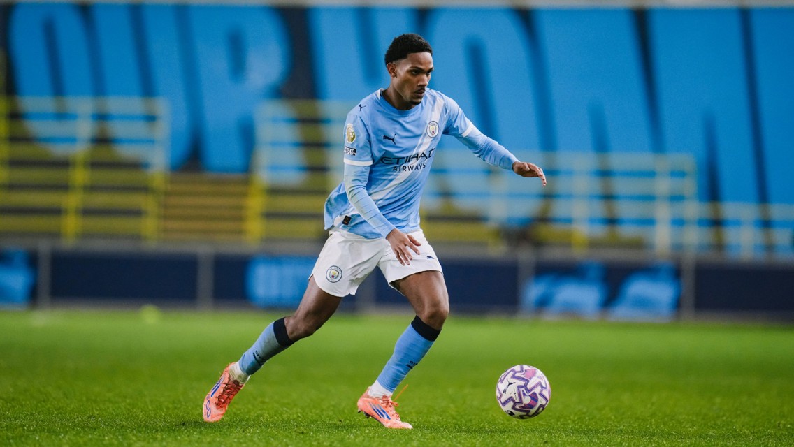 A Manchester City player dribbles a soccer ball on a field. The player is wearing the team's light blue kit and orange shoes. The background has blue and black signage.