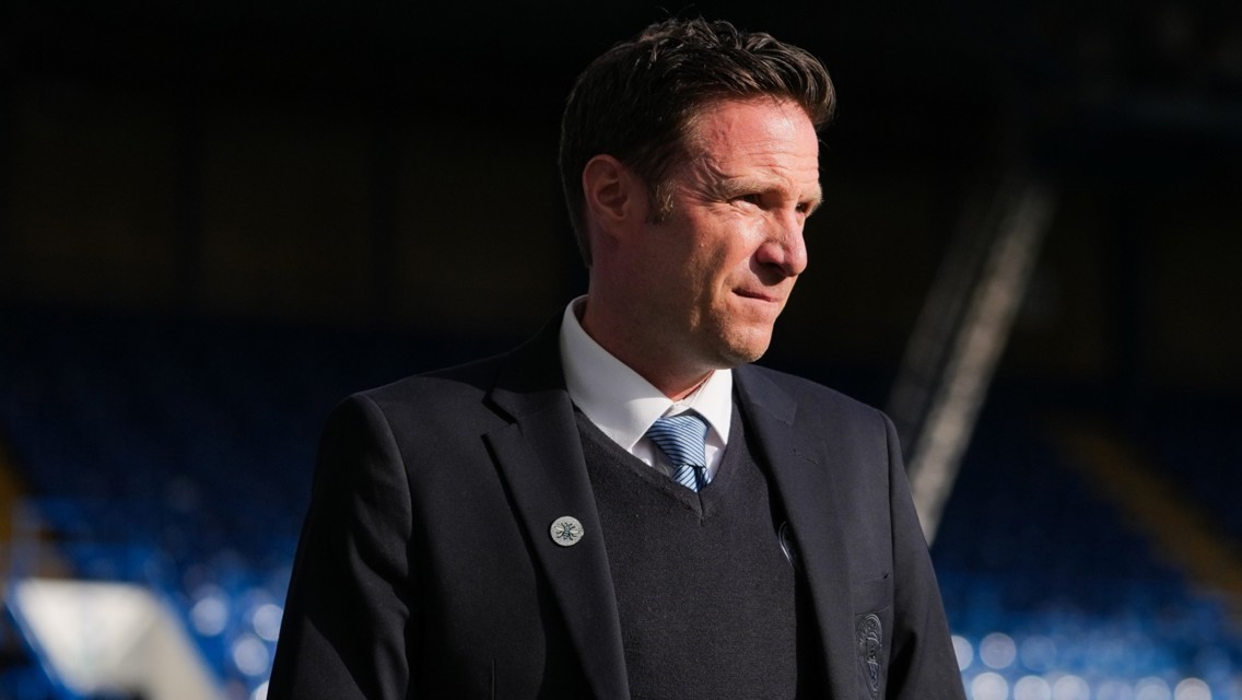 A person wearing a dark suit, sweater, and light blue tie, standing in a stadium.