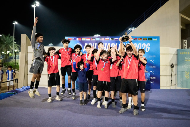 A youth football team celebrates winning a trophy at the Abu Dhabi Cup 2026. The players are wearing red and black uniforms and gold medals, and one player is lifting the trophy.