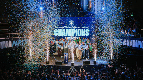 A group of people celebrating on stage with confetti falling. The backdrop reads 'Barclays Women's Super League Champions' and shows the year '25/26'.