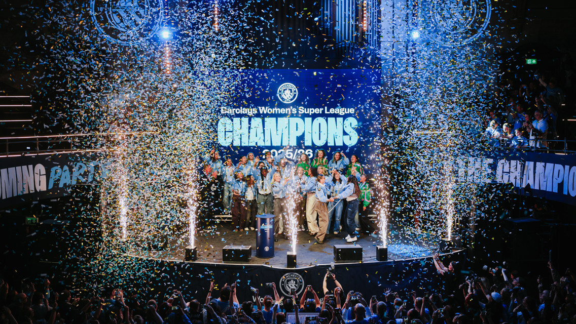 A group of people celebrating on stage with confetti falling. The backdrop reads 'Barclays Women's Super League Champions' and shows the year '25/26'.