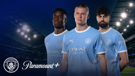 Three football players in Manchester City jerseys stand in front of a stadium backdrop with Manchester City and Paramount+ logos.