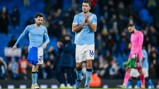 Players from Manchester City wearing light blue jerseys stand on the field after a match. The central figure has the number 16 on his jersey. The surrounding area shows a stadium with spectators in the background.