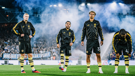 Four athletes in black tracksuits stand on a foggy stadium field, appearing focused.