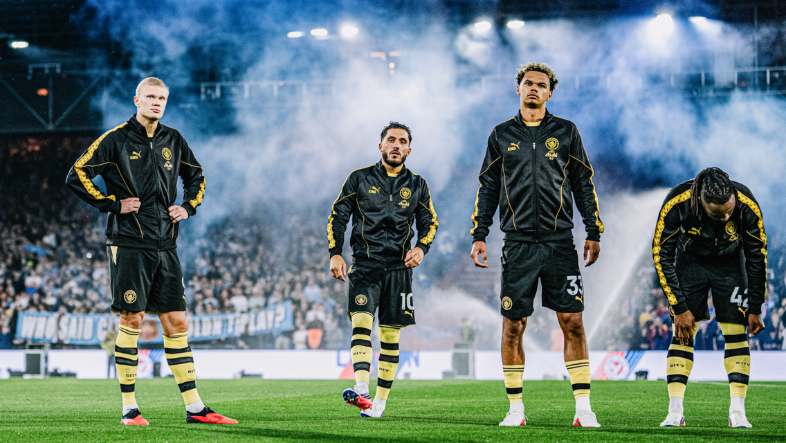 Four athletes in black tracksuits stand on a foggy stadium field, appearing focused.