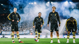 Four athletes in black tracksuits stand on a foggy stadium field, appearing focused.