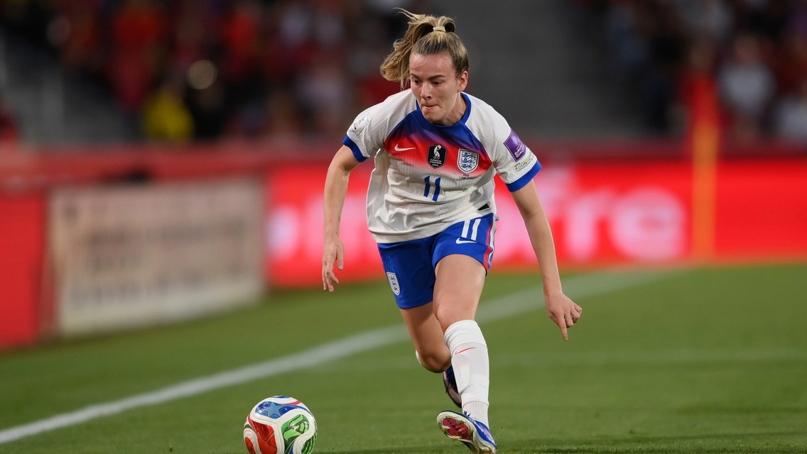 A female soccer player in an England jersey, number 11, dribbles the ball during a match. The uniform features blue shorts and socks with a white jersey and logos.