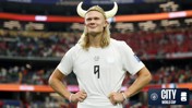 A football player wearing a white jersey stands with hands on hips, adorned with a Viking helmet. The stadium in the background has spectators and banners. The image includes a logo with text: 'CITY AT THE WORLD CUP'.