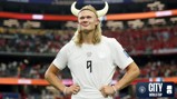A football player wearing a white jersey stands with hands on hips, adorned with a Viking helmet. The stadium in the background has spectators and banners. The image includes a logo with text: 'CITY AT THE WORLD CUP'.