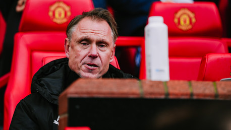 A person with a blurred face sits in front of red seats at a stadium with Manchester United logos.