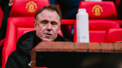 A person with a blurred face sits in front of red seats at a stadium with Manchester United logos.