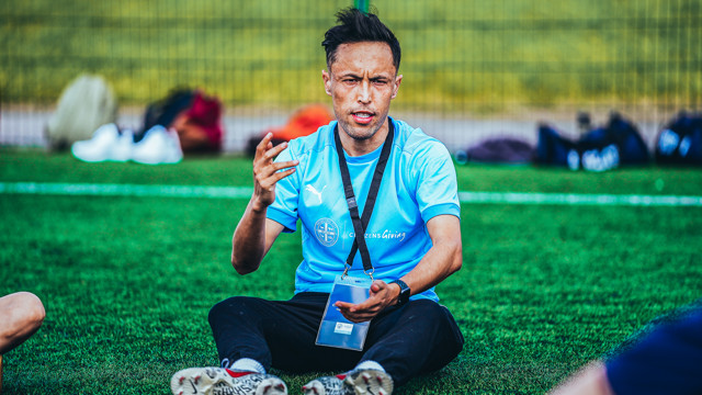 An individual in a light blue Manchester City shirt sits on a grassy field, engaged in a conversation, with blurred background objects suggesting a sports setting.