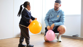 A child holding an orange balloon interacts with an adult wearing a Manchester City jacket holding a pink balloon.