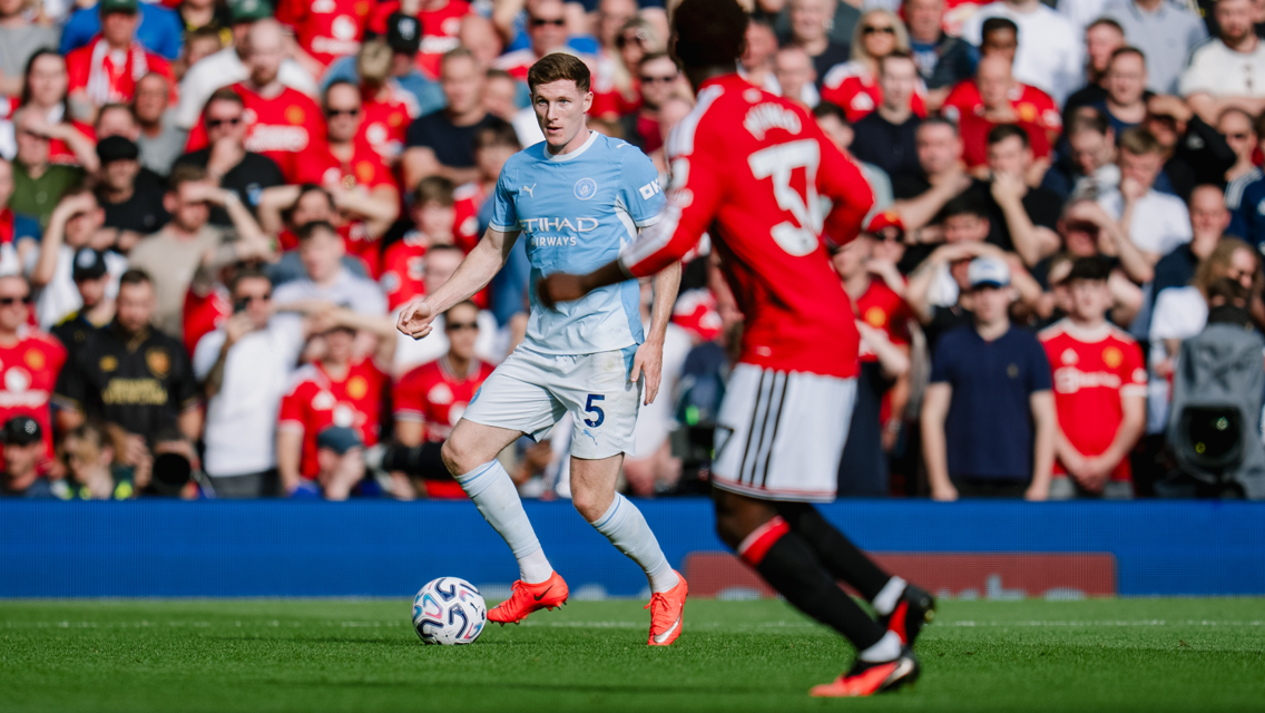 A football player from a team in light blue jersey controls the ball while an opponent in a red jersey approaches, with a crowd of fans in the background.