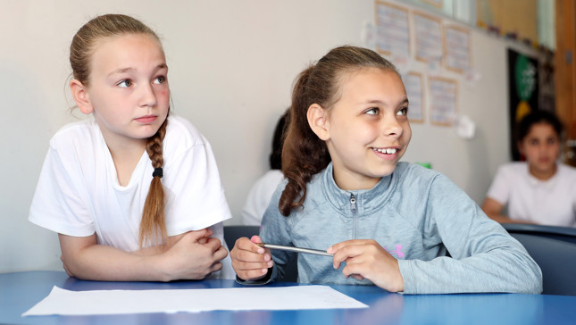 Two children sitting at a table in a classroom, one holding a pen and paper in front of them.