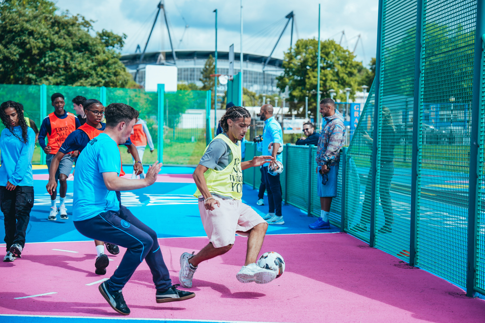 A group of people playing football on a colorful urban field with the Etihad Stadium in the background.