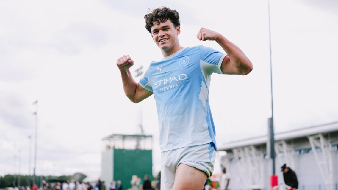 A football player in a light blue Manchester City kit is celebrating with raised fists on a field.