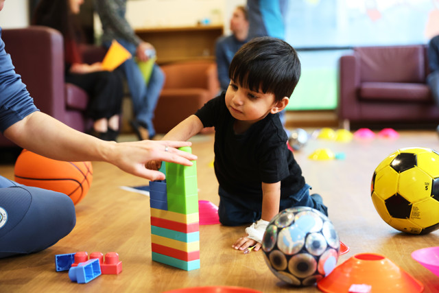 A child is playing with colorful toy blocks on a floor surrounded by balls and other toys, in a setting likely to be a playroom or a community center.
