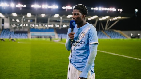 A Manchester City player is being interviewed on a football pitch at night. The player is wearing the team's sky blue kit and holding a microphone. The stadium lights are shining brightly.
